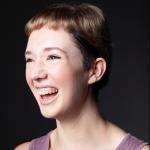 Phoebe Deklerk is looking to the right of the camera and laughing, she is wearing a mauve tank top. The background is almost black. 